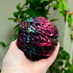 Pu&ograve; includere: Un gomitolo di lana con un mix di colori rosa, viola, blu e nero. La lana &egrave; lavorata a maglia a mano a forma di palla.
