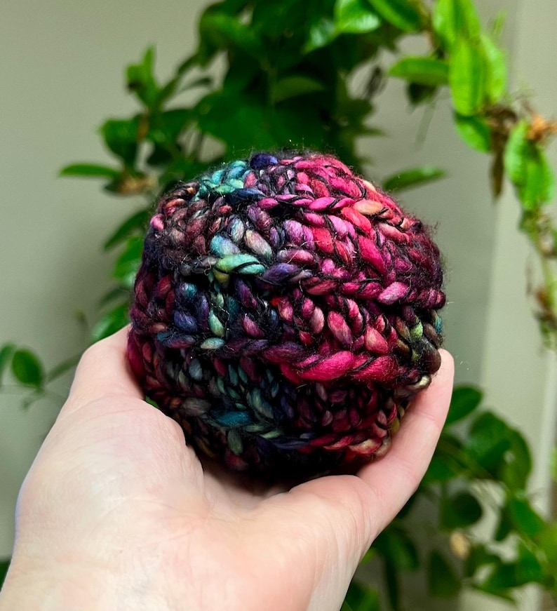 Pu&ograve; includere: Un gomitolo di lana con un mix di colori rosa, viola, blu e nero. La lana &egrave; lavorata a maglia a mano a forma di palla.