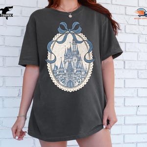 May include: Dark gray t-shirt featuring a detailed illustration of a fairytale castle in shades of blue. The castle is framed by a light blue ribbon and a decorative oval border. The shirt has a relaxed fit and short sleeves.