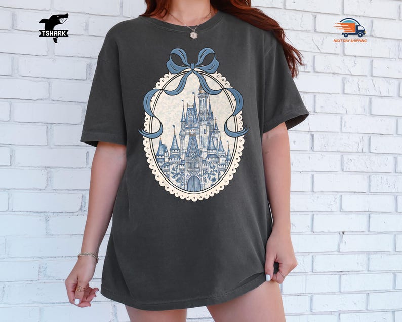 May include: Dark gray t-shirt featuring a detailed illustration of a fairytale castle in shades of blue. The castle is framed by a light blue ribbon and a decorative oval border. The shirt has a relaxed fit and short sleeves.