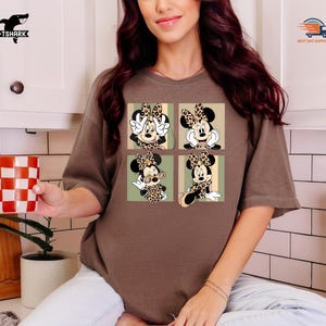 May include: Brown t-shirt featuring a four-panel design of Minnie Mouse in various poses. Each panel showcases Minnie with a leopard print bow and outfit, set against a light green and beige background. The shirt has short sleeves and a relaxed fit.
