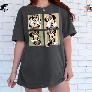 May include: Dark gray t-shirt featuring a four-panel graphic of Minnie Mouse in various poses, each with a leopard print bow and outfit. The shirt has a relaxed fit and a crew neck.