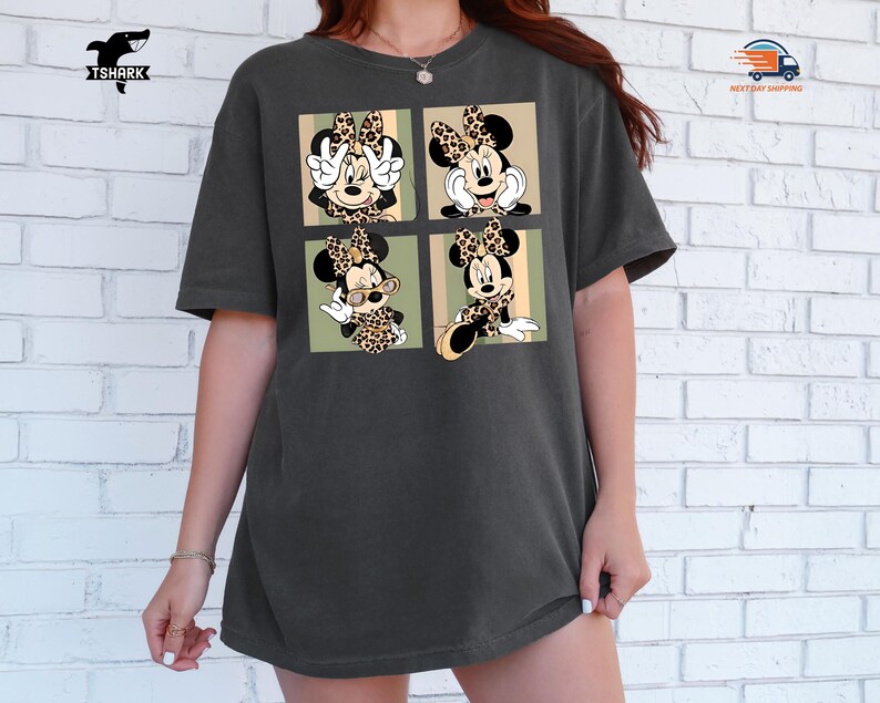 May include: Dark gray t-shirt featuring a four-panel graphic of Minnie Mouse in various poses, each with a leopard print bow and outfit. The shirt has a relaxed fit and a crew neck.