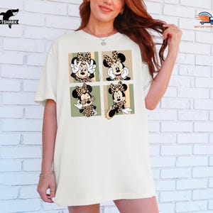 May include: Cream-colored t-shirt featuring a four-panel design of Minnie Mouse in various poses. Each panel showcases Minnie with a leopard print bow and accents. The shirt has a relaxed fit and short sleeves, perfect for casual wear.