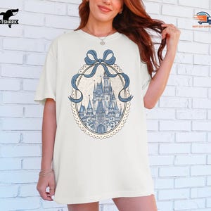 May include: Cream-colored t-shirt featuring a blue castle design within a decorative oval frame with a blue bow. The castle is detailed with towers and spires. The shirt has short sleeves and a relaxed fit.
