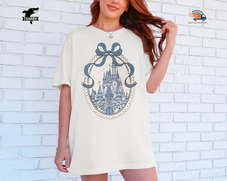 May include: Cream-colored t-shirt featuring a blue castle design within a decorative oval frame with a blue bow. The castle is detailed with towers and spires. The shirt has short sleeves and a relaxed fit.
