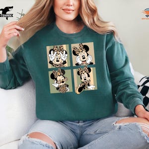 May include: Teal green sweatshirt featuring a four-panel design of Minnie Mouse in various poses. Each panel showcases Minnie with a leopard print bow and accents. The sweatshirt is a comfortable, casual style.