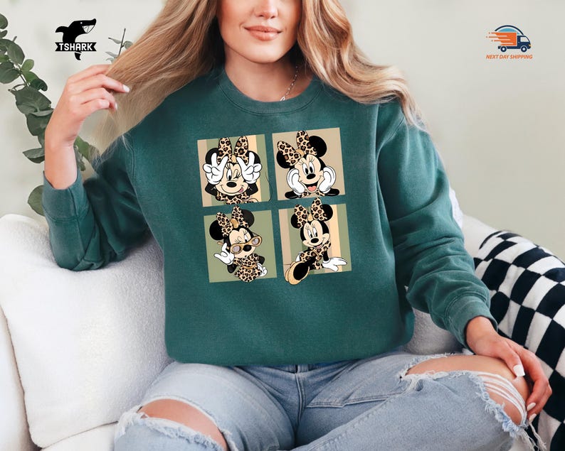 May include: Teal green sweatshirt featuring a four-panel design of Minnie Mouse in various poses. Each panel showcases Minnie with a leopard print bow and accents. The sweatshirt is a comfortable, casual style.