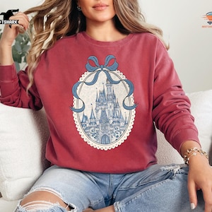 May include: A maroon sweatshirt featuring a detailed illustration of a fairytale castle within an oval frame, adorned with a blue ribbon. The castle is depicted in shades of blue and white, set against a cream-colored background.