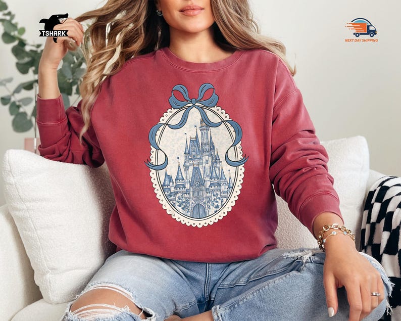 May include: A maroon sweatshirt featuring a detailed illustration of a fairytale castle within an oval frame, adorned with a blue ribbon. The castle is depicted in shades of blue and white, set against a cream-colored background.