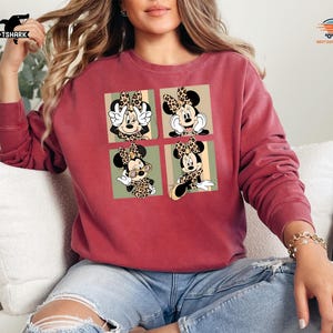 May include: A maroon sweatshirt featuring a four-panel design of Minnie Mouse in various poses, each with a leopard print bow and accents. The sweatshirt has long sleeves and a relaxed fit, perfect for casual wear.