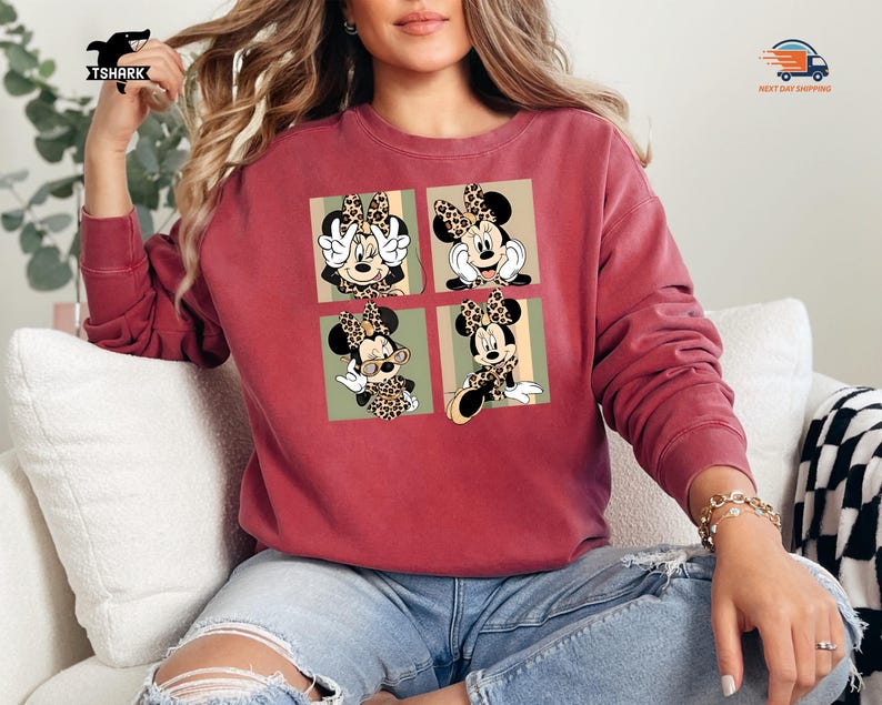 May include: A maroon sweatshirt featuring a four-panel design of Minnie Mouse in various poses, each with a leopard print bow and accents. The sweatshirt has long sleeves and a relaxed fit, perfect for casual wear.