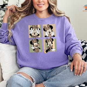 May include: A light purple sweatshirt featuring a four-panel design of Minnie Mouse in various poses, each with a leopard print bow. The sweatshirt is a solid color, and the design is in a square format.