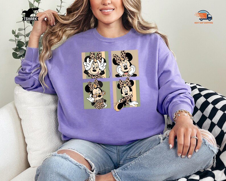 May include: A light purple sweatshirt featuring a four-panel design of Minnie Mouse in various poses, each with a leopard print bow. The sweatshirt is a solid color, and the design is in a square format.