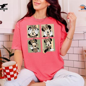May include: A coral-colored t-shirt featuring a four-panel design of Minnie Mouse in various poses, each with a leopard print bow and accents. The shirt is worn by a person holding a red and white checkered mug.