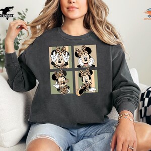 May include: Dark gray sweatshirt featuring a four-panel design of Minnie Mouse in various poses, each wearing a leopard print bow. The sweatshirt has a crew neck and long sleeves. The TSHARK logo is visible in the upper left corner.