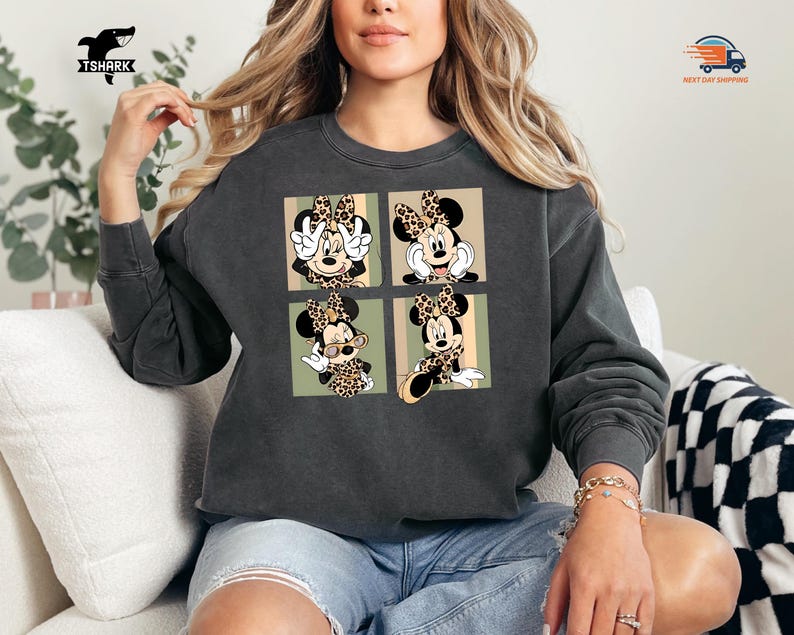 May include: Dark gray sweatshirt featuring a four-panel design of Minnie Mouse in various poses, each wearing a leopard print bow. The sweatshirt has a crew neck and long sleeves. The TSHARK logo is visible in the upper left corner.