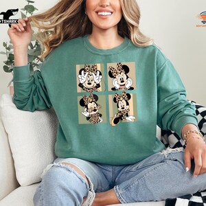 May include: A teal green sweatshirt featuring a four-panel design of Minnie Mouse in various poses. Each panel showcases Minnie with a leopard print bow and accents. The sweatshirt is a comfortable, casual style.
