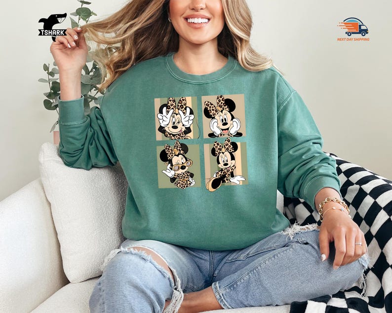 May include: A teal green sweatshirt featuring a four-panel design of Minnie Mouse in various poses. Each panel showcases Minnie with a leopard print bow and accents. The sweatshirt is a comfortable, casual style.
