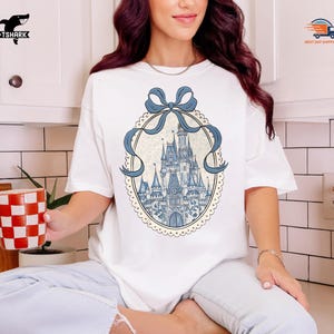 May include: White t-shirt featuring a blue and white castle design within an oval frame, adorned with a blue bow. The design is centered on the shirt. The shirt is a casual, everyday wear item.