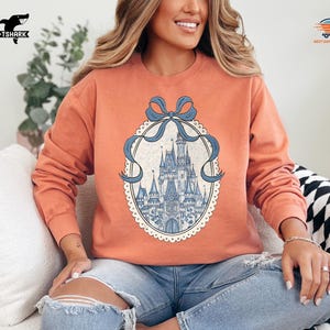 May include: Coral-colored sweatshirt featuring a detailed illustration of a castle within an oval frame, adorned with a blue bow. The castle design is in shades of blue and white. The sweatshirt has long sleeves and a crew neck.