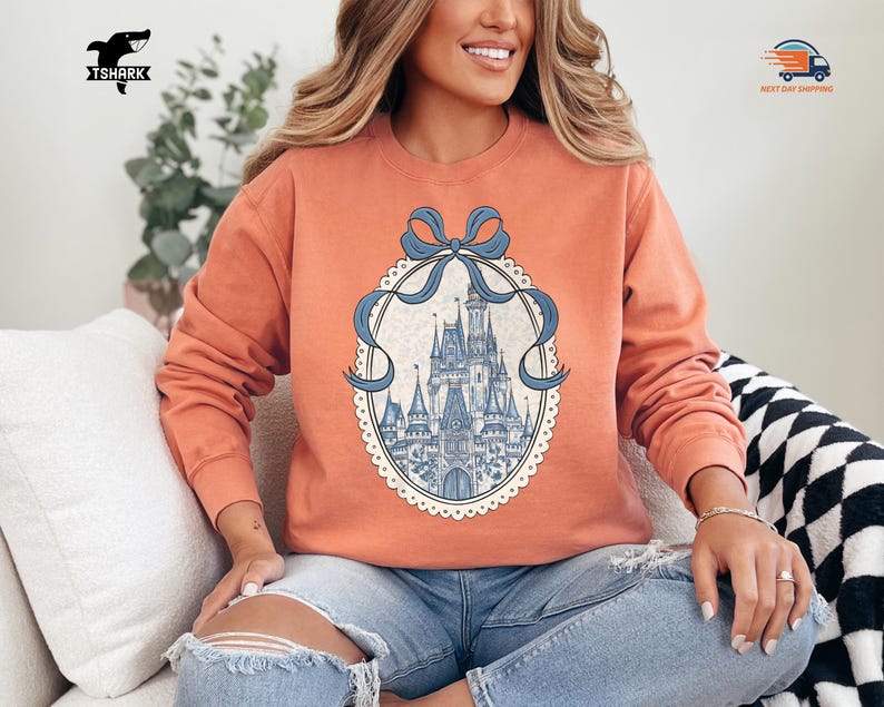 May include: Coral-colored sweatshirt featuring a detailed illustration of a castle within an oval frame, adorned with a blue bow. The castle design is in shades of blue and white. The sweatshirt has long sleeves and a crew neck.