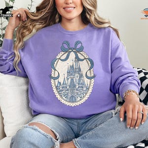 May include: A light purple sweatshirt featuring a detailed illustration of a castle within an oval frame, adorned with a blue ribbon. The sweatshirt is a solid color, and the castle illustration has blue and white accents.