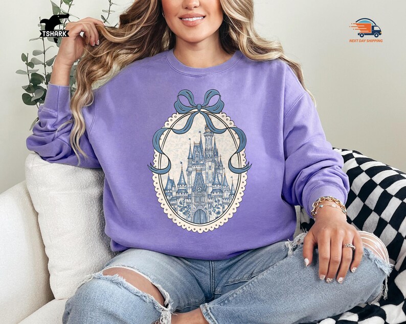 May include: A light purple sweatshirt featuring a detailed illustration of a castle within an oval frame, adorned with a blue ribbon. The sweatshirt is a solid color, and the castle illustration has blue and white accents.