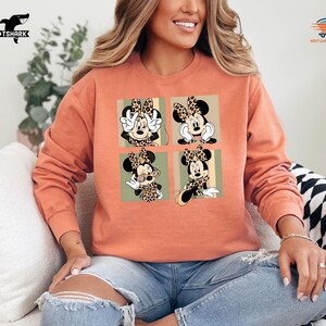 May include: A coral-colored sweatshirt featuring a four-panel design of Minnie Mouse in various poses. Each panel showcases Minnie with a leopard print bow and accents. The sweatshirt has a crew neck and long sleeves, perfect for casual wear.