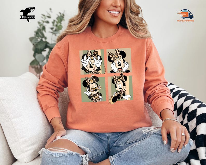 May include: A coral-colored sweatshirt featuring a four-panel design of Minnie Mouse in various poses. Each panel showcases Minnie with a leopard print bow and accents. The sweatshirt has a crew neck and long sleeves, perfect for casual wear.