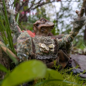 May include: A handcrafted puppet resembling a lizard-like being, featuring a textured brown face and body. It is dressed in a textured green and brown outfit adorned with circular embellishments. The puppet is positioned outdoors amidst natural green vegetation.