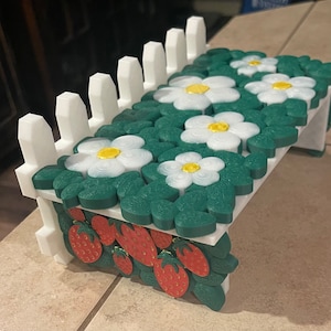 Op de afbeelding: Een wit hek omringt een miniatuurtuin met groene bladeren en witte bloemen met gouden centra. De tuin is gemaakt van plastic en is een decoratief item.