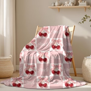 Puede incluir: Una manta a cuadros rosa y blanca con un patrón repetido de cerezas atadas con lazos rosas. El nombre "Ameli" está impreso en la manta. La manta está sobre una silla de madera.