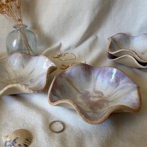 May include: Three ceramic bowls with wavy, scalloped edges and a glossy finish featuring a blend of soft lavender, white, and cream colors. The bowls have a smooth interior surface and a natural, unglazed tan exterior base.