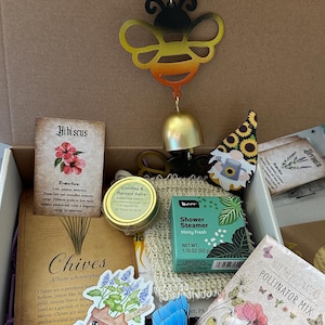 May include: A gift box filled with various items. Includes a metal bee wind chime, a jar of salve, a shower steamer, a lip balm, a butterfly decoration, and seed packets. There are also cards with text about herbs and flowers.
