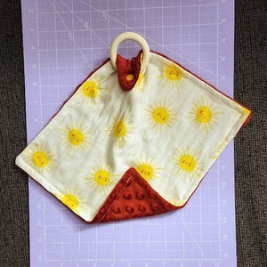 May include: A red and white baby sensory toy with a wooden ring attached. The white side features a pattern of yellow suns with smiling faces. The red side is a soft, textured fabric.