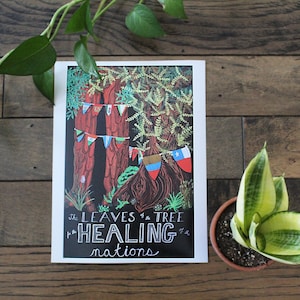 May include: A print featuring a detailed illustration of trees with colorful flags and the text "The Leaves of the Tree Healing Nations." The artwork is set against a dark background and framed in white, with a plant in a pot on the right.