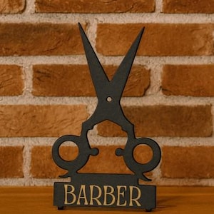 Puede incluir: Letrero de barbería de metal negro con un par de tijeras estilizadas. La palabra "BARBER" se muestra en letras doradas sobre una base rectangular. El letrero está colocado contra una pared de ladrillos.