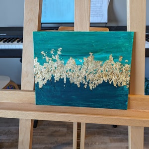 May include: A teal and gold abstract painting on canvas. The painting features a horizontal band of gold leaf across the middle of the canvas, with teal paint above and below.