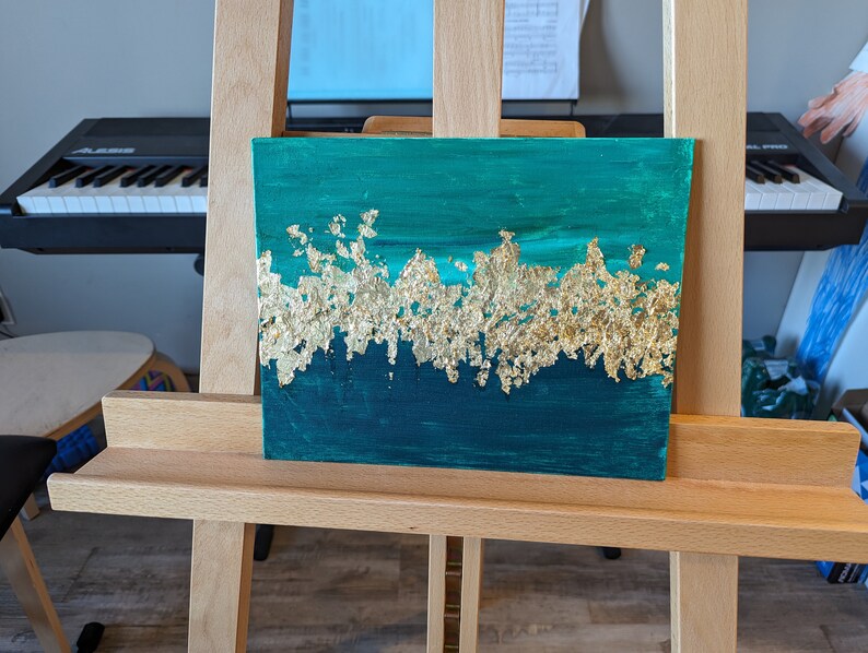 May include: A teal and gold abstract painting on canvas. The painting features a horizontal band of gold leaf across the middle of the canvas, with teal paint above and below.