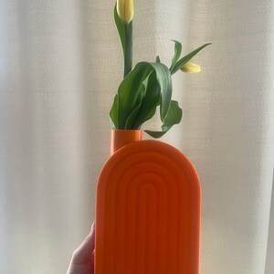 May include: Orange ceramic vase with a rounded arch design. The vase is filled with yellow tulips and green leaves.
