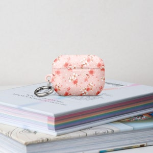 May include: A pink Airpods case with a floral pattern and a silver keychain. The case is sitting on top of a stack of books.