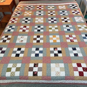May include: A patchwork quilt featuring a repeating nine-patch block pattern. The design incorporates various fabric squares in shades of red, blue, yellow, green, and white, separated by muted green and pink textured sashing strips.