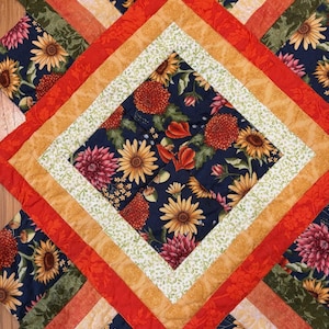 May include: A quilted textile featuring a diamond pattern. The center diamond showcases a dark blue floral print with sunflowers and dahlias. Surrounding layers include orange, gold, and green fabrics, creating a vibrant, geometric design. The quilt is likely handmade.