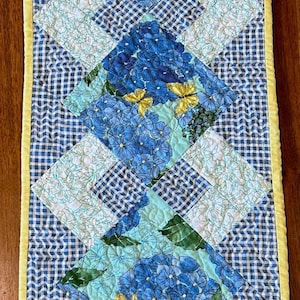 May include: A blue and white gingham table runner with a yellow border and a floral pattern featuring blue flowers and yellow butterflies.