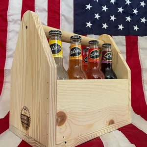 May include: A wooden six-pack carrier with four glass bottles of Mike's Hard Lemonade. The bottles are in the colours red, orange, yellow, and purple. The carrier is sitting on a red, white and blue American flag.