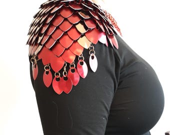 May include: A shoulder armour piece constructed from overlapping metallic red scales in a layered pattern. The lower edge features individual dangling red scales attached by small metal rings, creating a fringe effect over a plain black long-sleeve shirt.