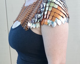 May include: A silver and copper chainmail shoulder piece with a decorative design. The shoulder piece is made of small metal scales and is worn over a black vest top.