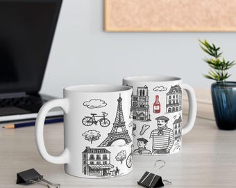 May include: Two white ceramic mugs with black line drawings of Parisian landmarks and icons. The Eiffel Tower, a bicycle, and a cafe are among the illustrations. One mug is in front of the other, with a laptop and office supplies in the background.