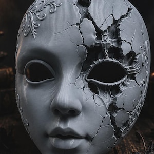 May include: A grey, human-like face mask featuring intricate, raised floral scrollwork patterns on the forehead and sides. The right side of the mask displays a prominent, jagged crack pattern that extends across the eye socket and cheek area.